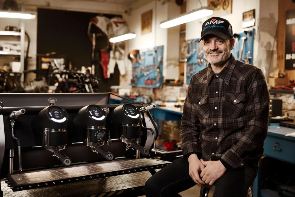 Hombre sentado junto a una cafetera profesional de espresso en un taller con herramientas de fondo